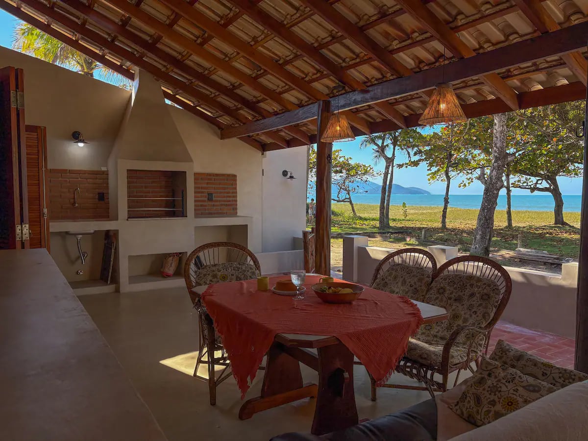 Varanda coberta da Casa Tohmé reformada, com mesa de jantar e vista para o mar entre as árvores.
