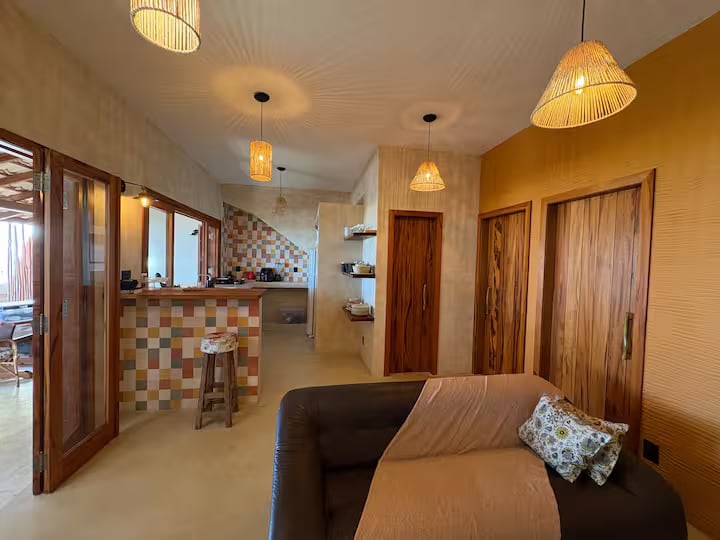Sala da Casa Tohmé depois da reforma, com sofá, luminárias de palha, cozinha ao fundo e portas de madeira.